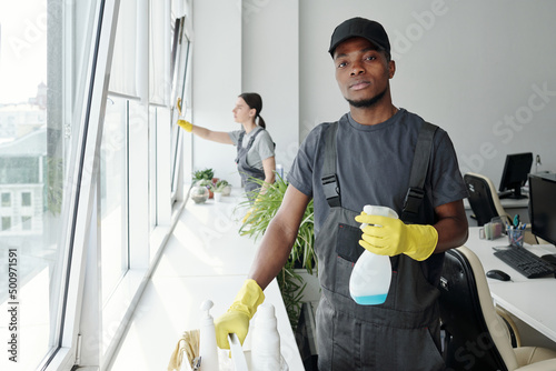 Young black man in coveralls, t-shirt, cap and rubber gloves using detergent in plastic sprayer while standing by window in office