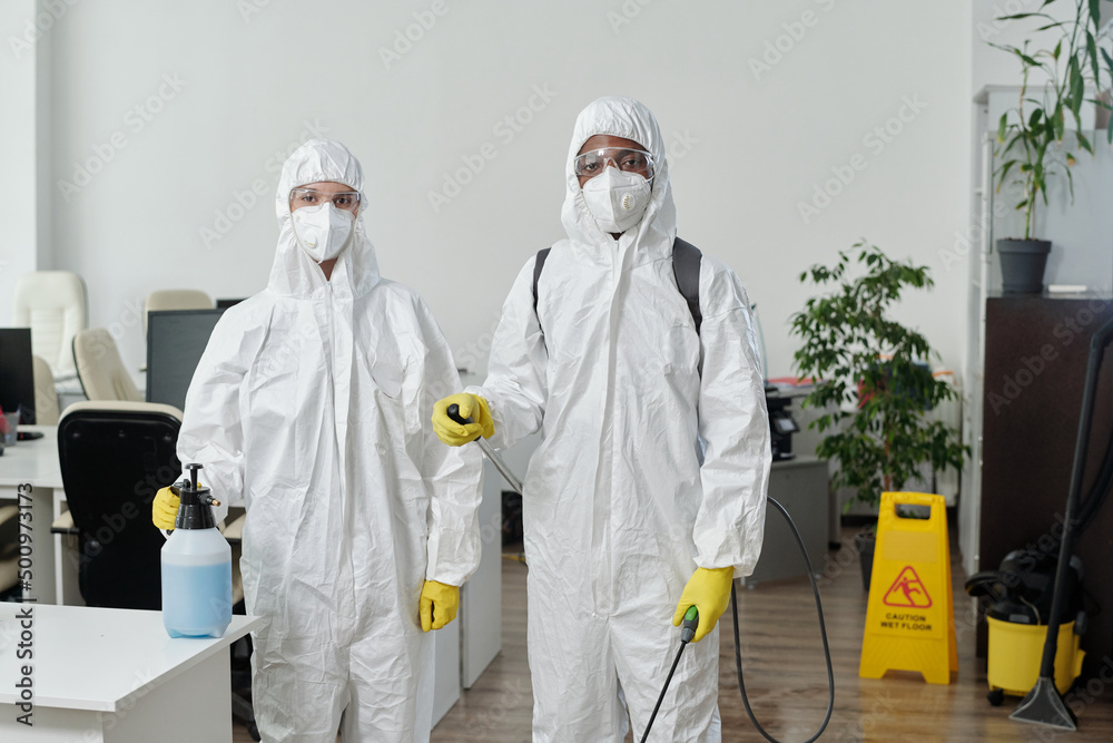 Small group of cleaning service company workers in hazmat suits looking at camera while spraying disinfectant all around openspace office Small group of cleaning service company workers in hazmat suits looking at camera while spraying disinfectant all around openspace office