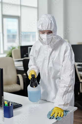 Contemporary young cleaning service worker with sprayer disinfecting desks and wiping their surfaces in large openspace office photo