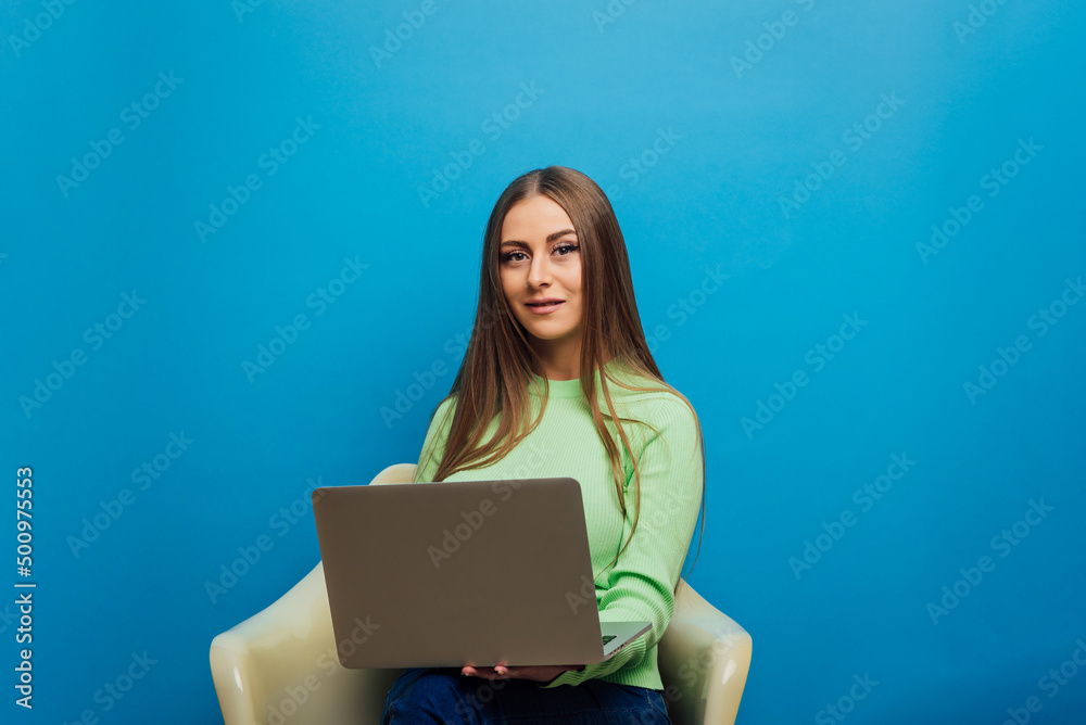 Woman looking at camera while using a laptop computer sitting on a chair over an isolated background. Technology and business concept. Woman looking at camera while using a laptop computer sitting on a chair over an isolated background. Technology and business concept.