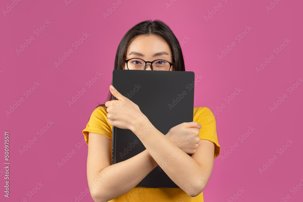 Asian female student in glasses hiding behind laptop, studying for college exams online, standing on violet background Asian female student in glasses hiding behind laptop, studying for college exams online, standing on violet background