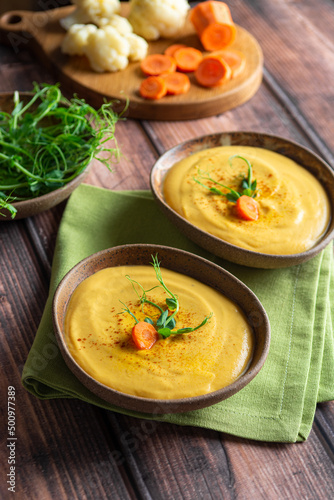 Vegetarian carrot and cauliflower cream-soup garnished with microgreens in ceramic bowls on a wooden table with chopped vegetables on the background. Healthy food concept.