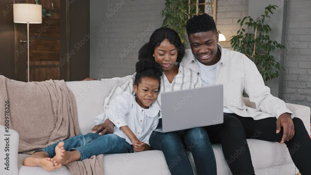Happy African family and cute kid child girl daughter using laptop looking at computer screen enjoying watching funny social media video having online video call conference relaxing on sofa at home