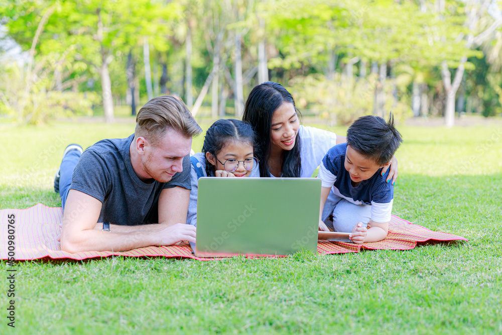 Happy family are lying on picnic blanket, they were enjoying talking about their summer vacation plan with their son and daughter using through the laptop in nature park. Family vacation background. Happy family are lying on picnic blanket, they were enjoying talking about their summer vacation plan with their son and daughter using through the laptop in nature park. Family vacation background.