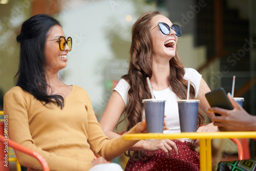 Young women in singlasses joking around and laughing when meeting in outdoor cafe on Suturday afternoon photo