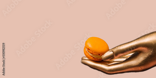 A golden female hand holds an orange macaroon, on a dirty pink background photo