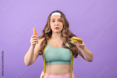 Serious hungry young european plus size woman with carrot and burger in hands, makes choice