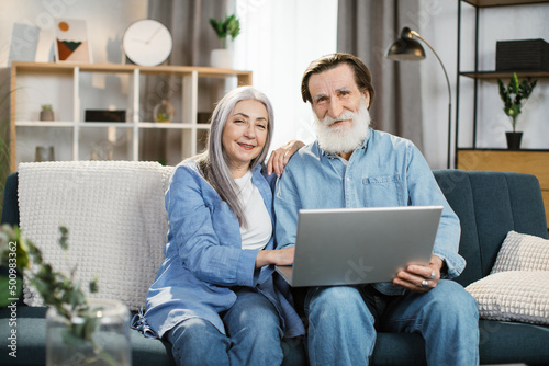 Happy mature couple Using Laptop Watching Movie Together Sitting On Couch At Home. Older Couple Browsing Internet On Computer Reading Online News On Weekend. Technology And Gadgets