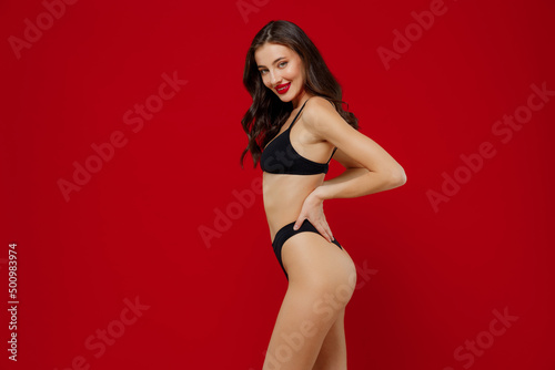 Side profile view young sexy smiling brunette woman 20s with perfect fit body in black underwear hold hand on waist stand akimbo isolated on plain red background studio . People female beauty concept