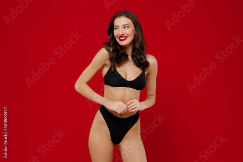 Young sexy shy smiling fun woman 20s with perfect fit body wear black underwear look aside on workspace area copy space isolated on plain red background studio portrait. People female beauty concept