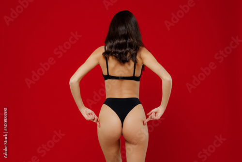 Back view young sexy brunette woman with perfect fit body in black underwear point finger on buttocks without cellulite isolated on plain red background studio portrait. People female beauty concept