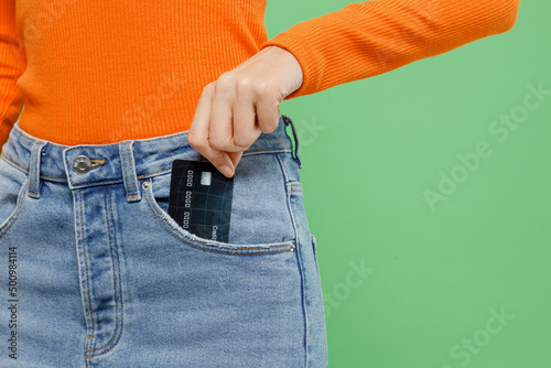 Cropped close up woman 20s wear casual orange short hold in hand put credit bank card to denim pocket isolated on plain pastel light green color background studio portrait. People lifestyle concept.