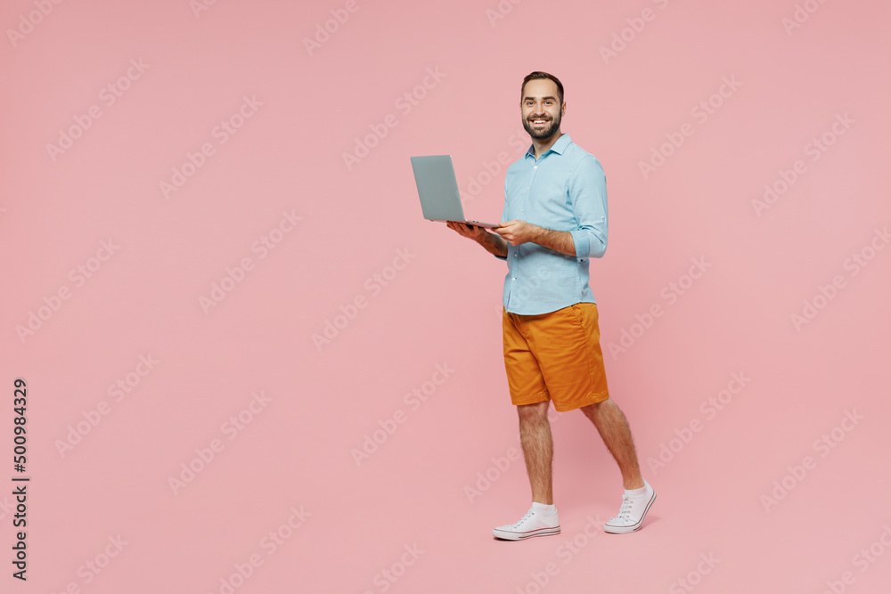 Full body side view freelancer young smiling man 20s in classic blue shirt hold use work on laptop pc computer isolated on plain pastel light pink background studio portrait. People lifestyle concept. Full body side view freelancer young smiling man 20s in classic blue shirt hold use work on laptop pc computer isolated on plain pastel light pink background studio portrait. People lifestyle concept.