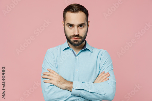 Young strict sad serious caucasian man 20s wearing classic blue shirt look camera holding hands crossed folded isolated on plain pastel light pink background studio portrait. People lifestyle concept.