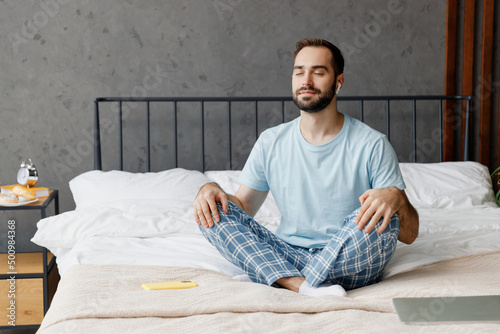 Young peaceful man in casual blue t-shirt sitting on bed in lotus pose yoga om aum gesture relax meditate try to calm down listen music in earphones rest relax spend time in bedroom home in own room. photo