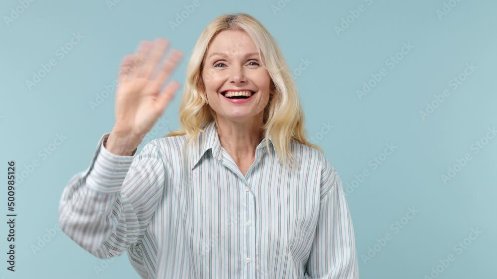 Fun elderly blonde woman lady 40s years old wears white shirt look around for friend find waving meet greet with hand as notices someone isolated on plain pastel light blue background studio portrait Fun elderly blonde woman lady 40s years old wears white shirt look around for friend find waving meet greet with hand as notices someone isolated on plain pastel light blue background studio portrait