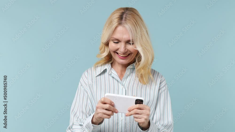 Gambling elderly blonde woman lady 40s years old wears white shirt using play racing on mobile cell phone hold gadget for pc video games isolated on plain pastel light blue background studio portrait Gambling elderly blonde woman lady 40s years old wears white shirt using play racing on mobile cell phone hold gadget for pc video games isolated on plain pastel light blue background studio portrait
