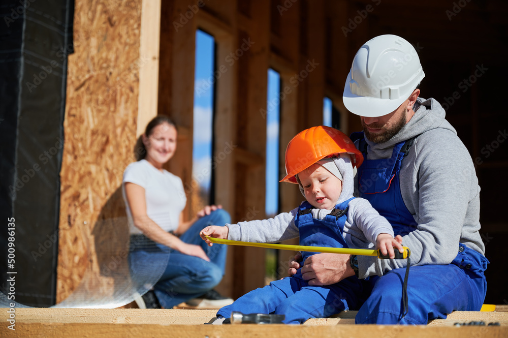Father, mother and son building wooden frame house. Toddler boy helping his daddy, while woman looking for them on construction site. Guys wearing helmet and blue overalls. Carpentry, family concept. Father, mother and son building wooden frame house. Toddler boy helping his daddy, while woman looking for them on construction site. Guys wearing helmet and blue overalls. Carpentry, family concept.