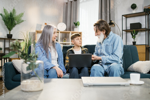 Happy mature couple and their handsome grandson boy relax together in living room, watching video or browsing internet using modern computer device. Concept of family leisure, teaching of retirees.