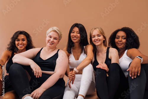 Five smiling women in fitness clothes sitting together on pastel background. Females with different body types relaxing after workout.