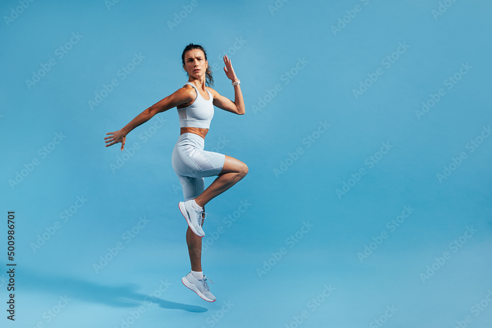 Slim woman jumping in studio on blue background. Muscular female in fitness clothes exercising.