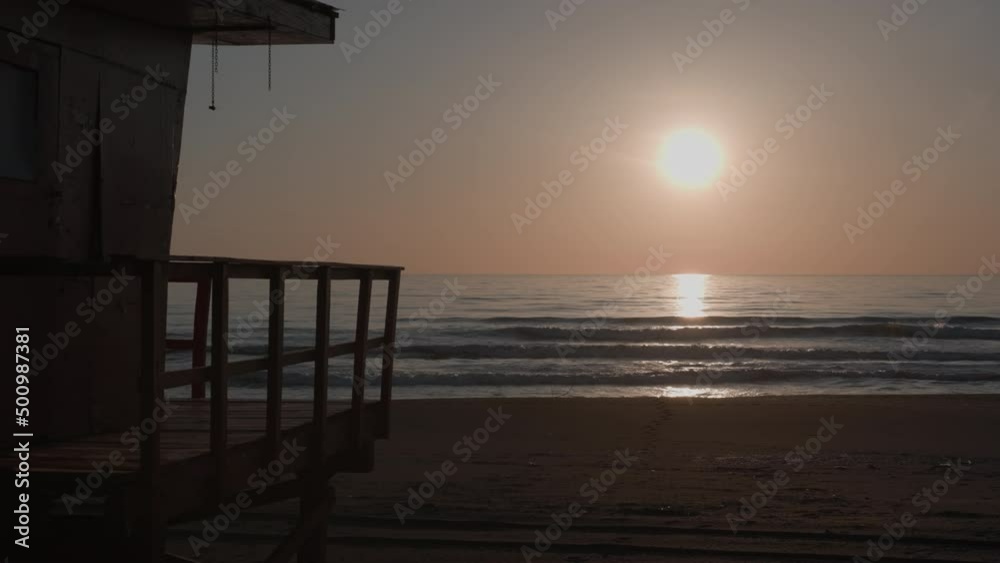 Lifeguard tower silhouette at sunset time on empty beach with moderate waves. Beautiful tranquil slow motion of sea at evening time.