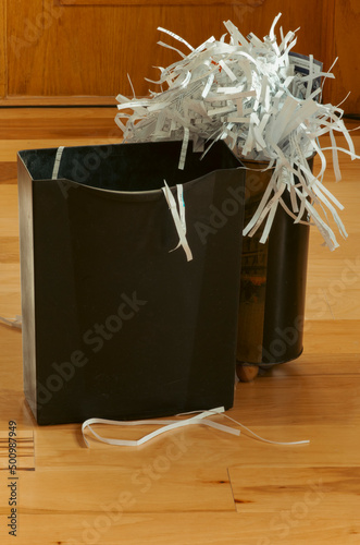 Shredded paper overflows wastebasket with another empty basket near. Office space is wood flooring and walls signifying a home office setting. Closeup of shred and baskets.