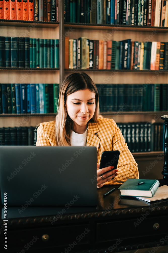 Portrait of young woman college student using mobile phone, preparing for test exam, writing essay doing homework in the library. Portrait of young woman college student using mobile phone, preparing for test exam, writing essay doing homework in the library.