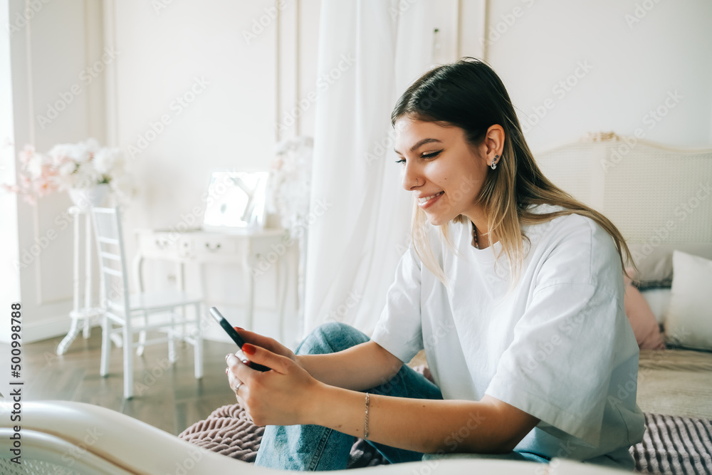 Portrait of young cheerful woman using mobile phone, sitting on a bed and looking on a screen. Portrait of young cheerful woman using mobile phone, sitting on a bed and looking on a screen.