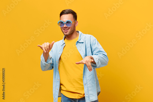 Fototapeta Overjoyed tanned handsome man in blue shirt trendy sunglasses dance sing posing isolated on orange yellow studio background