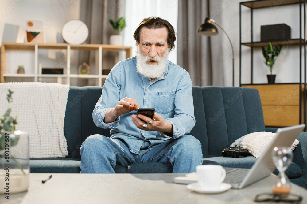Video call distance meeting social networks concept. Senior man with surprised expression on his face sitting on sofa at home having video call using his smartphone talking and having fun with friends