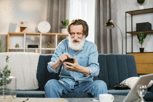 People home and using gadgets. Confident stylish man with grey beard smiling happily sitting on the couch at cozy living room indoors and using smartphone for typing message or serfing social networks