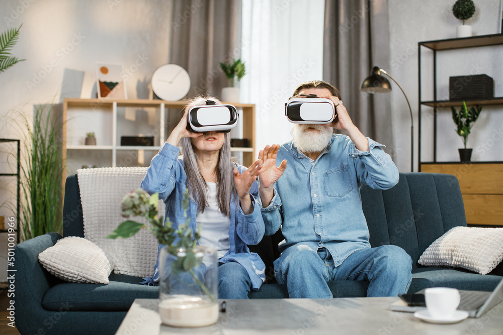 Happy mature family playing games at home and using VR glasses at home. Cheerful senior couple, family sitting on couch, sofa and entertaining themselves with innovations.