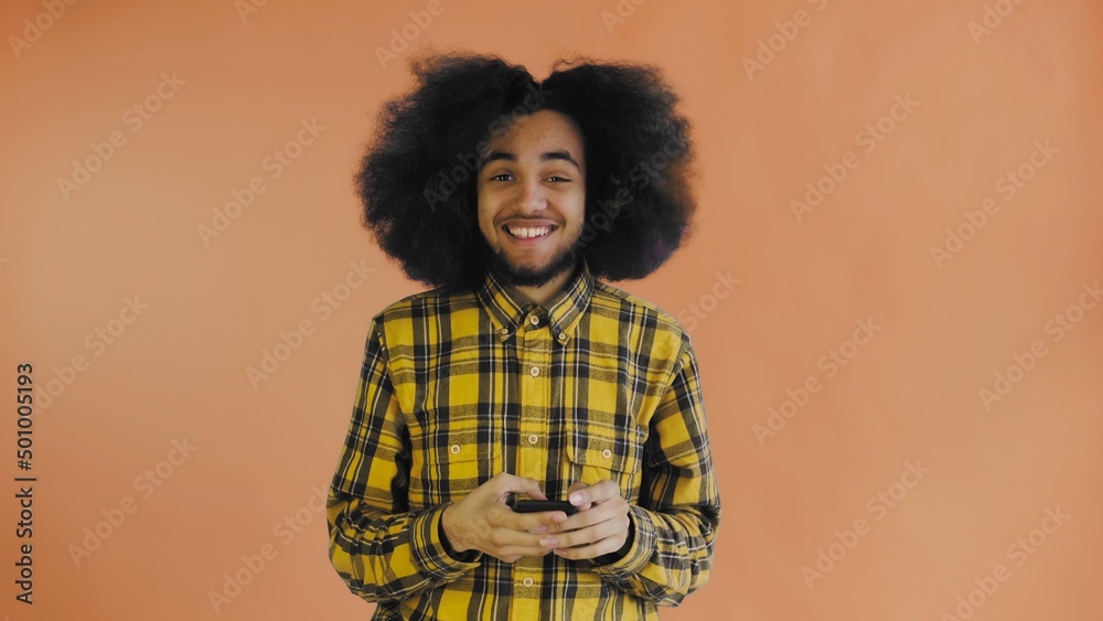 A young man with an African hairstyle on an orange background looks at the phone and is happily surprised. Emotions on a colored background A young man with an African hairstyle on an orange background looks at the phone and is happily surprised. Emotions on a colored background