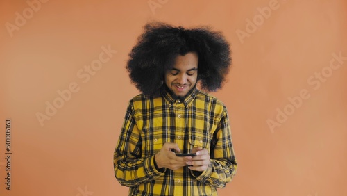 A young man with an African hairstyle on an orange background looks at the phone and happily texts with someone. Emotions on a colored background