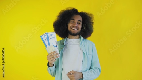 A happy African-American man looks at the camera, holds tourist vouchers in his hand, stands isolated on a yellow background
