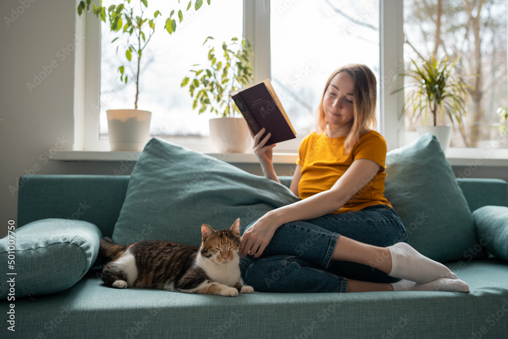 Young woman in yellow t-shirt reading book on sofa and petting cat. Young woman in yellow t-shirt reading book on sofa and petting cat.