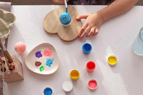 Kids hands paint an egg, on a white table with multi-colored paints in jars