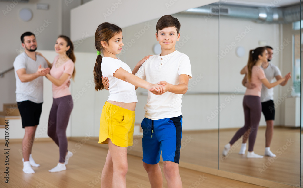 Pre-tee siblings practicing active dance in pair during family dance class, parents dancing on background