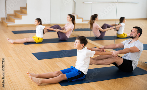 Smiling teenage boy exercising with father at couple yoga class while his mother practicing with his sister, practicing stretching in pairs