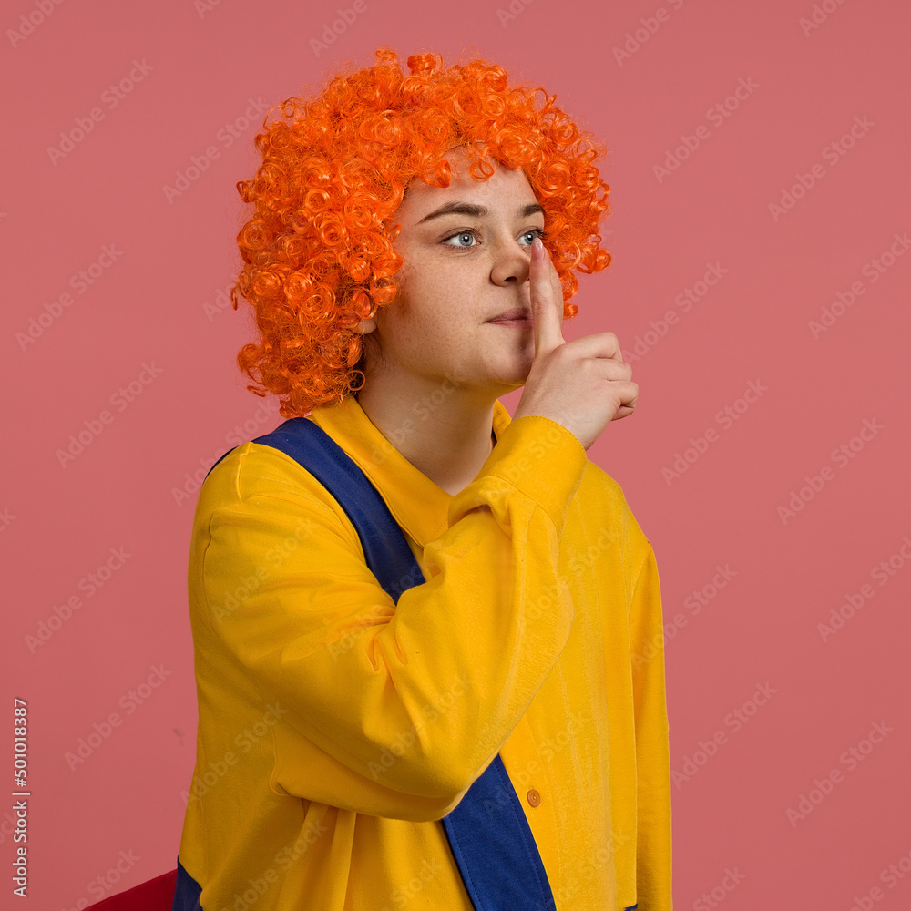a girl in a clown costume and a bright wig put one finger to her mouth, studio portrait on a colored background a girl in a clown costume and a bright wig put one finger to her mouth, studio portrait on a colored background