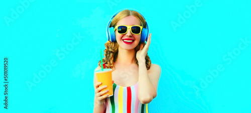 Portrait of happy smiling young woman listening to music in headphones with fresh cup of juice on blue background
