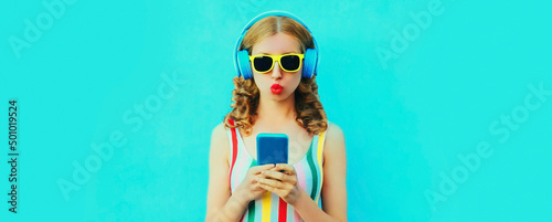 Portrait of stylish young woman listening to music in headphones with smartphone blowing her lips on blue background