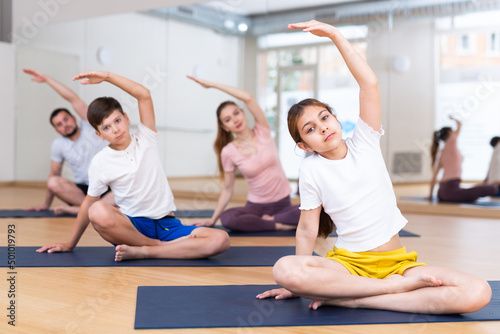 Cute tween girl with brother and parents exercising Hatha yoga in modern yoga studio, doing stretching in Padmasana pose..