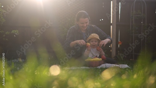 Happy Young Cheerful Mother Holding Baby Eating Fruits On Green Grass. Mom Adorable Infant Child Playing Outdoors With Love In Backyard Garden. Little Kid With Parents. Family, Nature, Ecology Concept