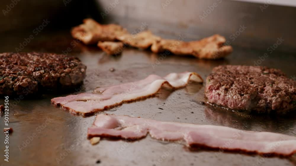 Close up of uncooked bacon and cooked burger in hot griddle and smoke coming out