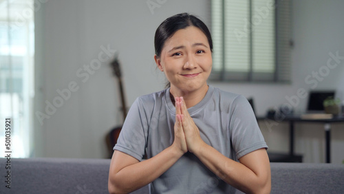Fotografia Asian woman holding hands in prayer, looking at camera sitting on sofa at home