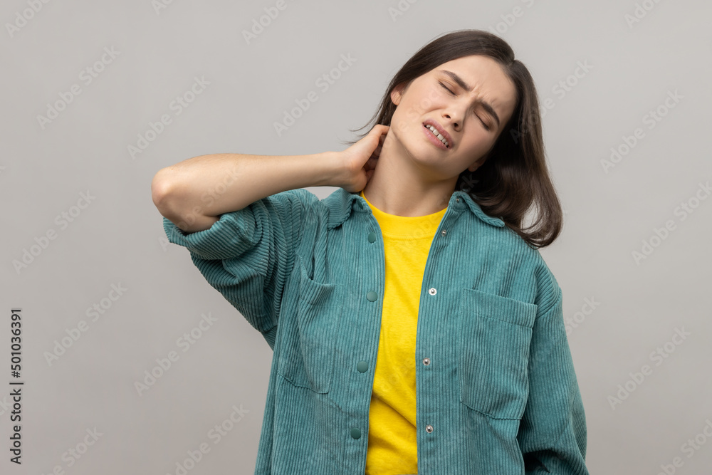 Unhealthy dark haired woman touching neck feeling pain and numbness, worried about muscle tension, osteochondrosis, wearing casual style jacket. Indoor studio shot isolated on gray background. Unhealthy dark haired woman touching neck feeling pain and numbness, worried about muscle tension, osteochondrosis, wearing casual style jacket. Indoor studio shot isolated on gray background.