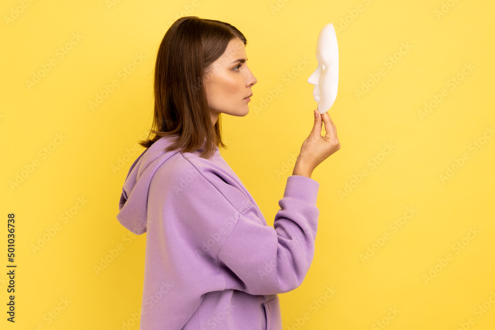 Profile of young adult attractive woman holding white mask in hands, hidden personality, expressing calm strict emotions, wearing purple hoodie. Indoor studio shot isolated on yellow background. Profile of young adult attractive woman holding white mask in hands, hidden personality, expressing calm strict emotions, wearing purple hoodie. Indoor studio shot isolated on yellow background.