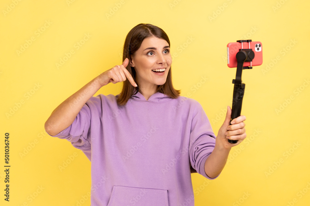 Portrait of woman using cell phone and steadicam for broadcasting livestream, pointing down, asking to subscribe, wearing purple hoodie. Indoor studio shot isolated on yellow background. Portrait of woman using cell phone and steadicam for broadcasting livestream, pointing down, asking to subscribe, wearing purple hoodie. Indoor studio shot isolated on yellow background.
